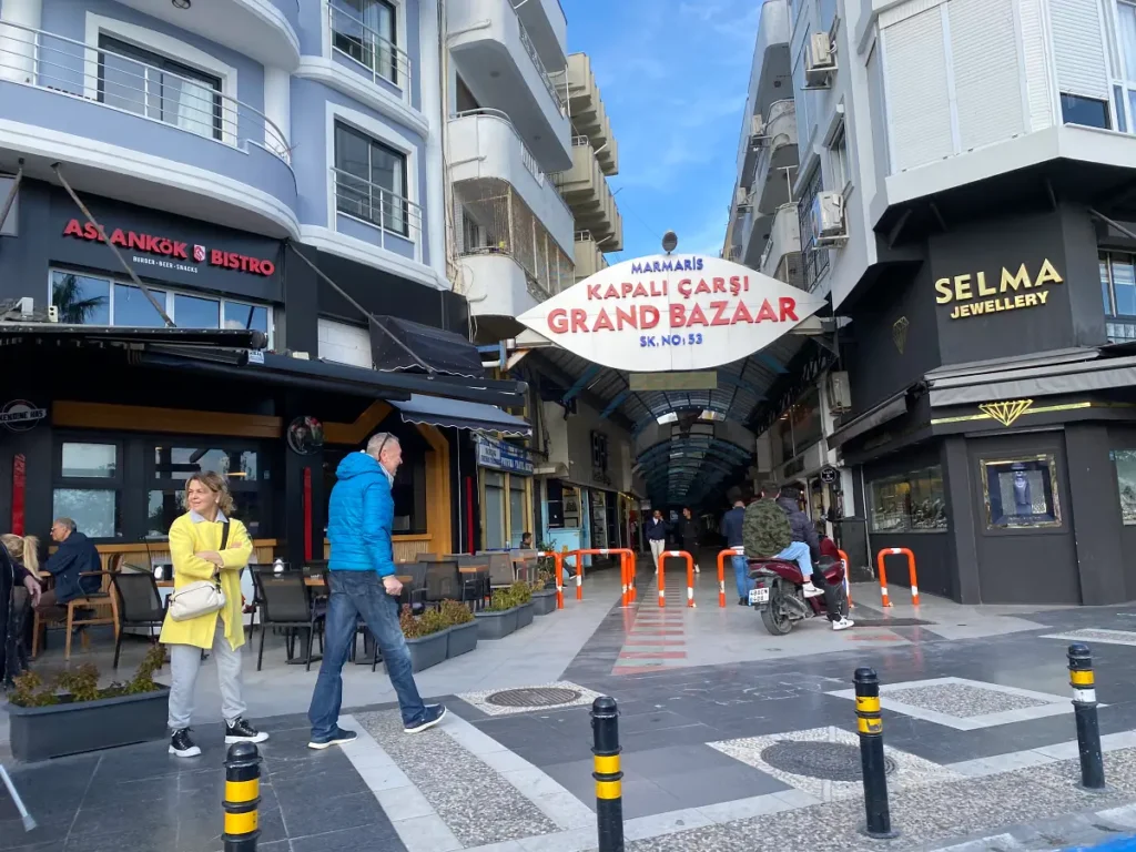Street-level view of the entrance to Marmaris Grand Bazaar in Turkey, with people walking by and shops lining the entrance, under a sign that reads "Marmaris Kapalı Çarşı Grand Bazaar