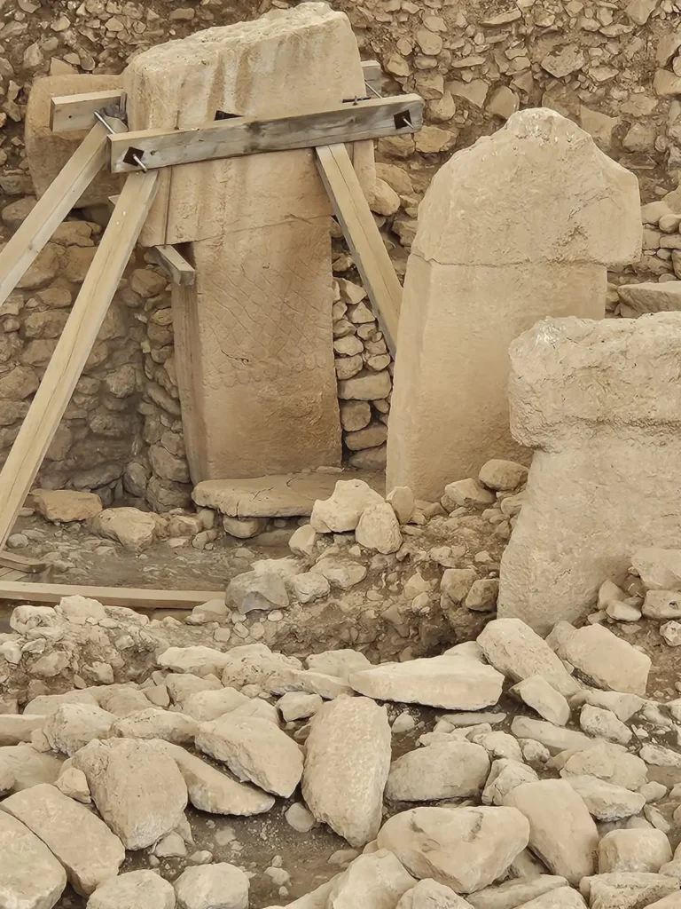 Photo of an archaeological site showcasing the ancient Göbekli Tepe. Visible are large, rounded limestone pillars, some standing upright with intricate carvings, and others lying horizontally amidst rubble. The pillars are supported by modern wooden braces, emphasizing the ongoing preservation efforts. The surrounding area is filled with smaller rocks and pebbles, with a backdrop of a partially excavated hillside, hinting at the extensive history still buried.