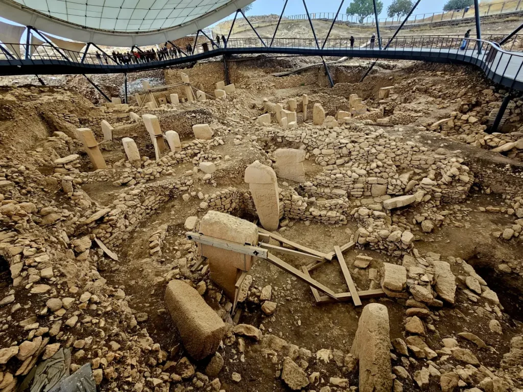 Wide-angle view of the Göbekli Tepe archaeological site. The photo shows a complex of unearthed T-shaped pillars of varying sizes, some upright and others fallen, amidst an array of ancient stone walls and floors. The excavation site is expansive with visible sections still under excavation, suggesting a grand scale of past human activity. Above, a modern canopy stretches across the site, while a metal walkway with visitors offers a glimpse into the ongoing interaction between the past and present. The image encapsulates the site's historical significance and the continuous efforts to preserve it.