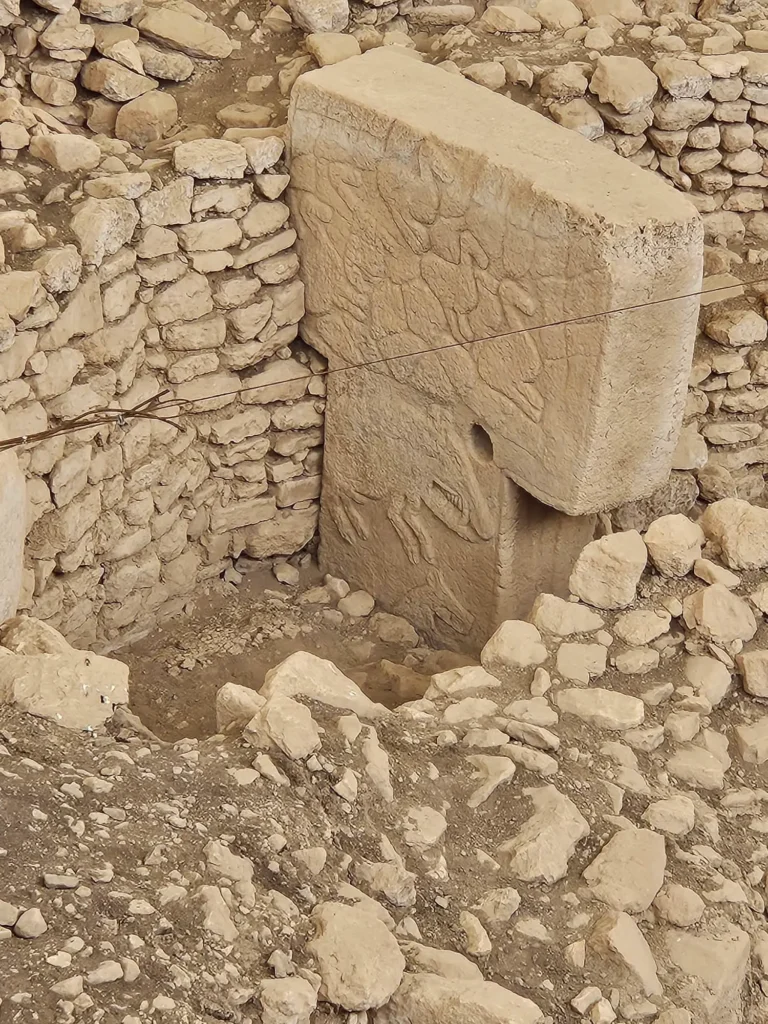 A close-up view of a large, rectangular limestone pillar at Göbekli Tepe, featuring a bas-relief carving of a reptile-like creature etched into the stone. The pillar, with its flat top and broad sides, stands out against the backdrop of smaller, rough-hewn stone blocks that appear to be part of an ancient wall. The carving is subtle yet visible, suggesting the significance of such depictions in the cultural or religious practices of the people who created it. The surrounding stones and earth provide context to the site's ancient and excavated nature.