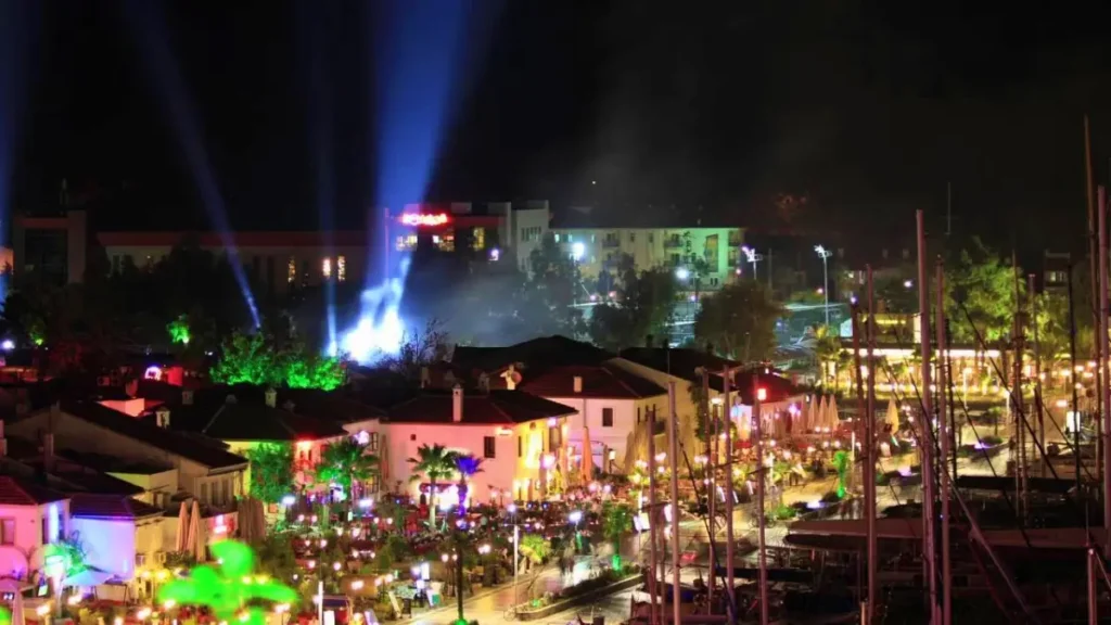 Nighttime view of a lively street in Marmaris, illuminated by colorful lights and lined with vibrant bars and restaurants, alive with activity and framed by moored boats and a dark sky pierced by beams from spotlights.