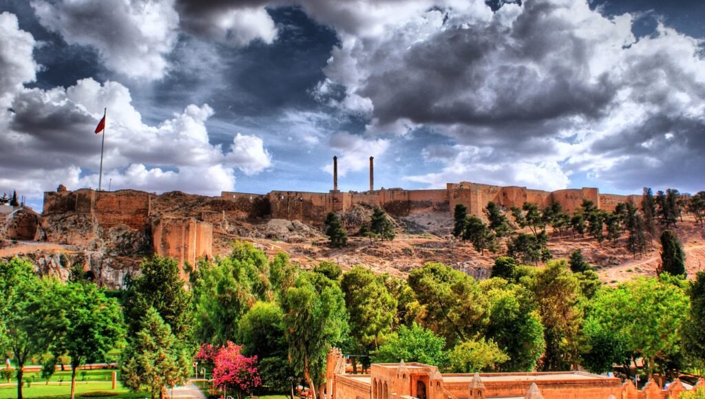 The photograph captures the imposing Sanliurfa Castle, perched on a hill and dominating the landscape. This ancient fortress, with its robust walls and distinctive twin columns, is a historical landmark in Sanliurfa, Turkey. The greenery surrounding the castle, including well-kept gardens and trees, provides a vibrant contrast to the stony architecture. Above, the sky is filled with expressive clouds, suggesting a lively yet serene atmosphere. The castle's strategic location and the flag flying high signify its historical importance and enduring presence in the region's cultural heritage.
