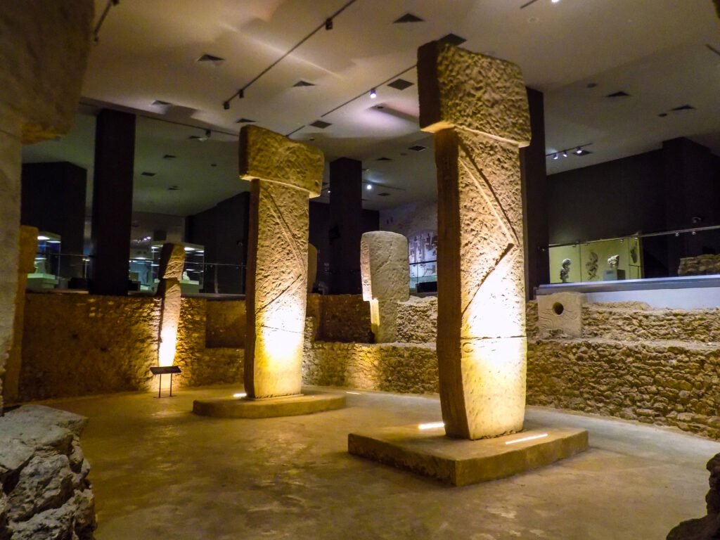 The image appears to show an indoor exhibit featuring the iconic T-shaped pillars from Göbeklitepe. These pillars are known for their prehistoric archaeological significance and are characterized by their abstract designs and animal carvings. The exhibit seems to be well-lit, highlighting the textures and carvings on the pillars, and it includes informational displays, likely providing context and history about Göbeklitepe. Such exhibits aim to bring ancient history to the public in a controlled environment, preserving the artifacts while allowing for education and appreciation of this significant archaeological site.