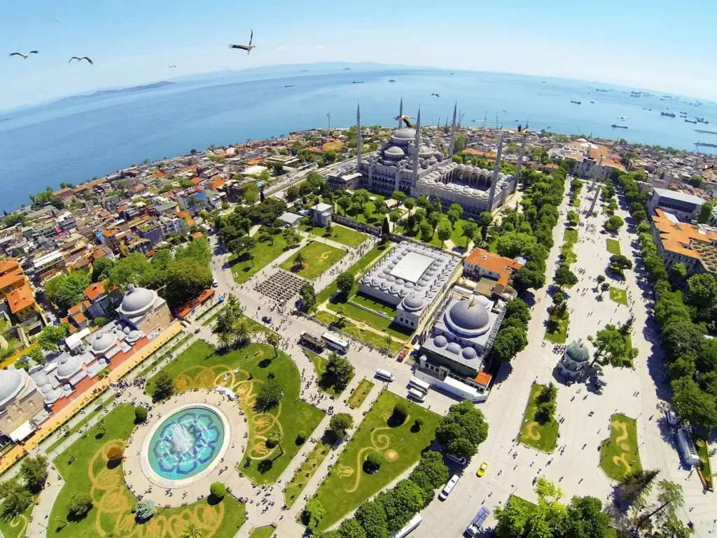 This aerial view presents a sweeping panorama of the historic Sultanahmet district in Istanbul, Turkey. Prominently featured are the Blue Mosque and Hagia Sophia, iconic landmarks of the city, set amid the lush greenery of Sultanahmet Park. The Marmara Sea glitters in the distance, dotted with ships, and the skies above are alive with seabirds, capturing the dynamic interplay between nature and the urban landscape.
