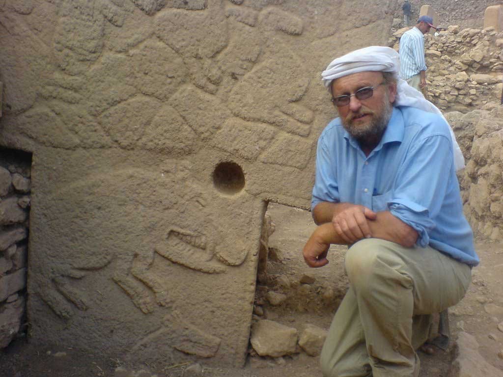 This image shows an Prof. Klaus Schmidt. sitting next to an archaeological stone structure with carvings. The person is dressed in a casual blue shirt and beige trousers, with a white headscarf. They appear to be resting one arm on their knee and the other on the stone structure while looking towards the camera. The stone has a circular hole and relief carvings that suggest it could be part of an ancient site. In the background, another person can be seen, possibly working on the site, indicative of an active archaeological dig. The image captures a moment of rest or contemplation amidst the exploration of history.