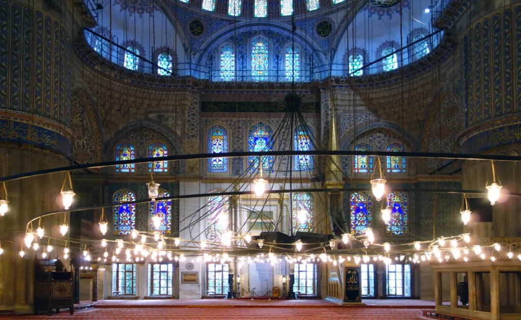 The interior shot of the Blue Mosque showcases the ornate detail and artistry that define the mosque's richly decorated spaces. The intricate designs of the Iznik tiles are visible, covering walls and archways in patterns of blue and turquoise, while the stained glass windows add vibrant splashes of color to the tranquil ambiance within. The lines of hanging lamps draw the eye towards the central prayer area, emphasizing the mosque's role as a place of worship and reflection. The architectural elements, such as the arches and the domes, are complemented by the elaborate calligraphy of Quranic verses that adorn the walls, resonating with the spiritual significance of the space. This image captures the essence of the Blue Mosque's beauty and serenity, inviting contemplation and appreciation for its historical and religious significance.