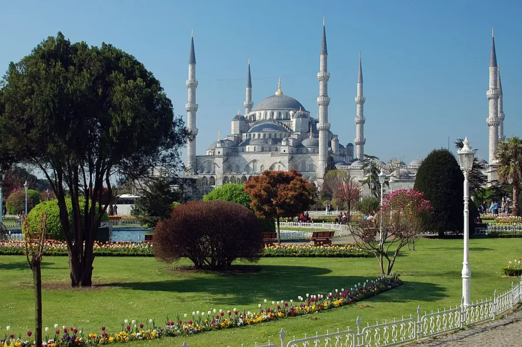 The image beautifully presents an external view of the Blue Mosque, also known as Sultan Ahmed Mosque, in Istanbul. The photo showcases the mosque's grand façade with its impressive array of minarets and domes, a signature of classic Ottoman architecture. The foreground is adorned with a well-manicured garden, adding a burst of color and life to the scene. The clear blue sky provides a perfect backdrop, emphasizing the mosque's silhouette. This view, often captured by visitors, represents the harmonious blend of nature and architecture, a serene setting inviting both contemplation and admiration. The Blue Mosque, in this setting, is not only a historical and religious symbol but also an integral part of the city's living landscape.