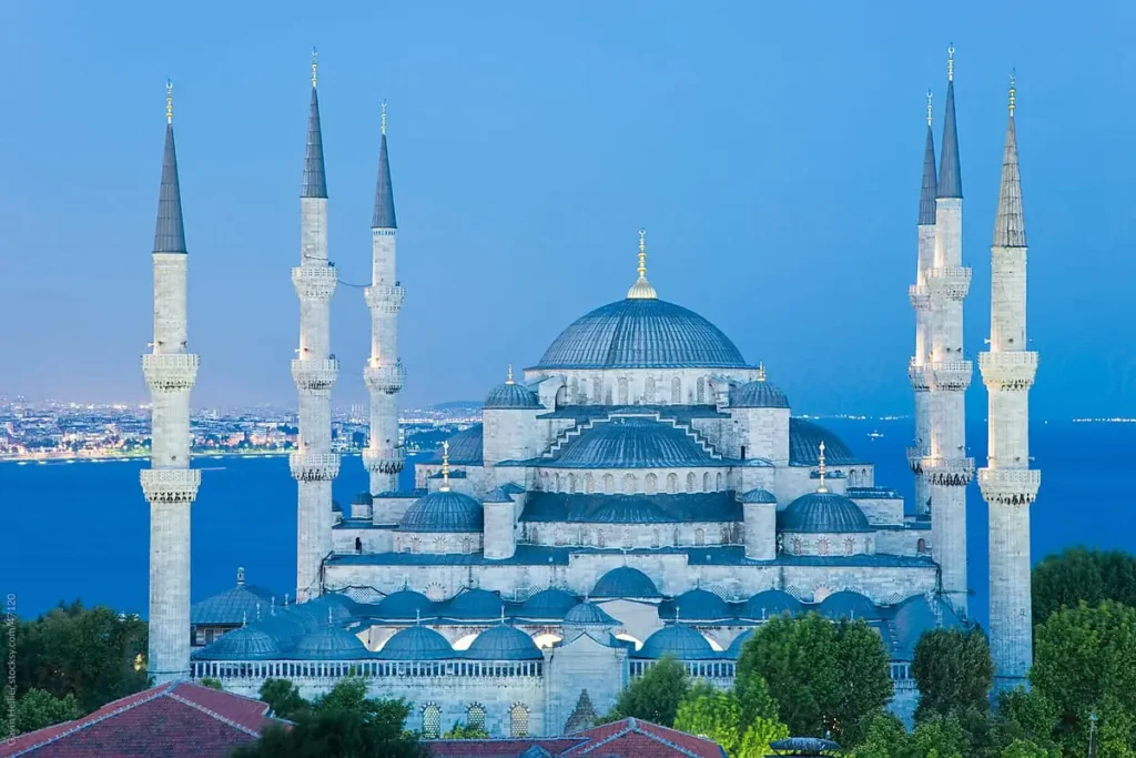 This is a stunning photograph of the Blue Mosque, also known as the Sultan Ahmed Mosque, located in Istanbul. The image captures the mosque in the evening light, with its distinctive minarets reaching towards the sky and the cascade of domes leading the eye to the grand central dome. The lighting accentuates the architectural details and highlights the mosque's symmetry and grandeur. The backdrop shows the beautiful cityscape of Istanbul and hints at the strategic location of the mosque, overlooking the Bosphorus Strait. This majestic sight is a testament to the mosque's status as both a historical monument and a centerpiece of Islamic architecture.