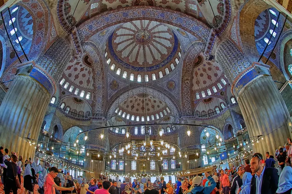 
ChatGPT
The photo you've shared captures the bustling atmosphere inside the Blue Mosque, with visitors and worshippers alike mingling beneath the grandeur of the mosque's interior. The image showcases the ornate patterns and designs that adorn the domes and the walls, as well as the series of hanging lights that add a warm glow to the space. The mosque's pillars rise majestically, supporting the vast domes, and the vibrant stained glass windows complement the blue of the tiles, creating a colorful display of light within the mosque. It's a vivid representation of the Blue Mosque as a place of both cultural convergence and religious devotion.