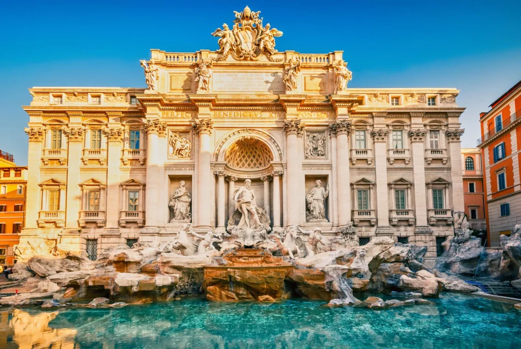 the magnificent Trevi Fountain in Rome during the day. The fountain is an architectural marvel, with sculptures of mythological figures set against a grand palace facade. Sunlight bathes the scene, highlighting the intricate details and the dynamic poses of the statues. The water in the fountain is a vibrant turquoise, providing a stark contrast to the warm tones of the travertine stone. The fountain's majestic design and scale dominate the frame, inviting the viewer to marvel at its Baroque splendor.