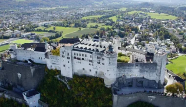 picture displays an aerial view of the Hohensalzburg Fortress, situated high above the cityscape of Salzburg. The surrounding landscape includes parts of the city and stretches of green fields, with a backdrop of mountains in the distance. The fortress and the city are bathed in bright daylight