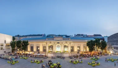 A wide-angle view of museumsquartier in Vienna during the evening. The image captures the lively atmosphere of the cultural district with people socializing on funky green seating arrangements. The classical facade of the Kunsthalle Wien is visible in the center, flanked by the contemporary designs of the Leopold Museum on the left and the mumok on the right. The square is vibrant with activity as visitors stroll, relax, and dine at open-air cafes under the fading light of the sky.